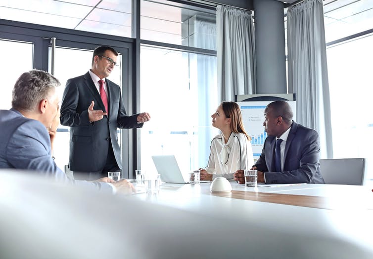 A businessman is standing at a table in a meeting room talking to a black man and woman.