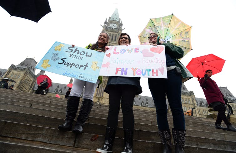 Three women stand together with signs that say 