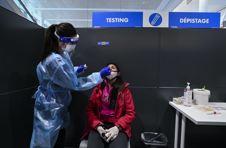 A woman in a red jacket receives a PCR test from a nurse wearing personal protective equipment.  A sign reading test is upon their heads.