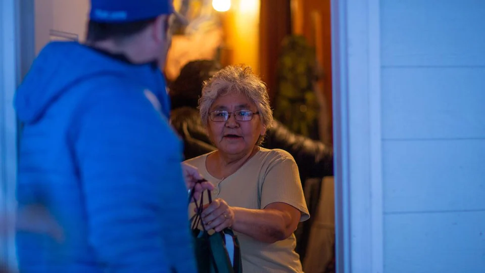 Un homme en bleu tend un sac à une femme qui se tient dans le cadre d'une porte.