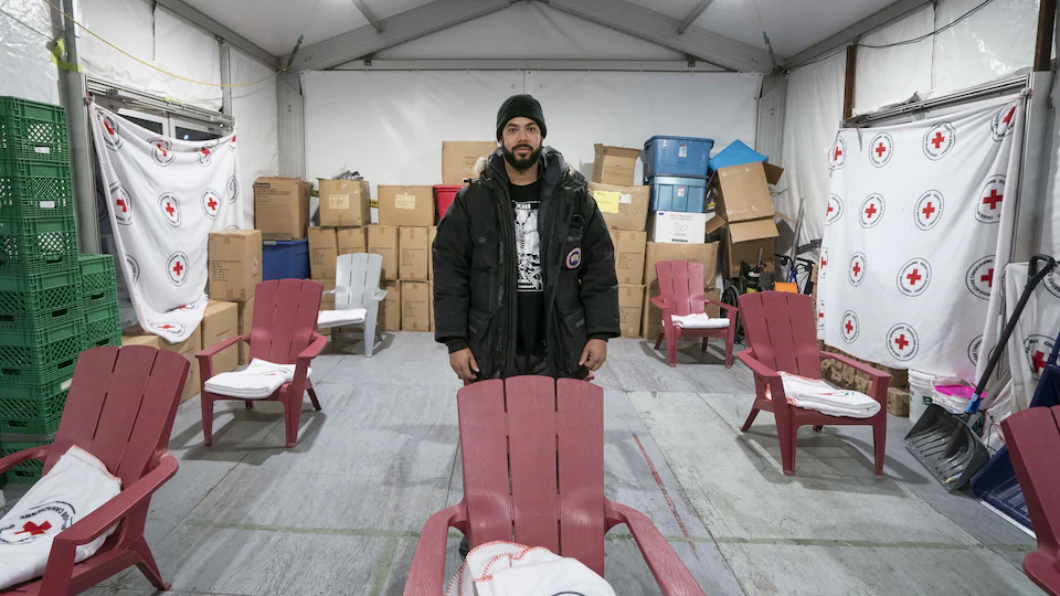 Un homme au milieu d'une pièce, devant une grande chaise rouge sur laquelle est posée une couverture de la Croix-Rouge.