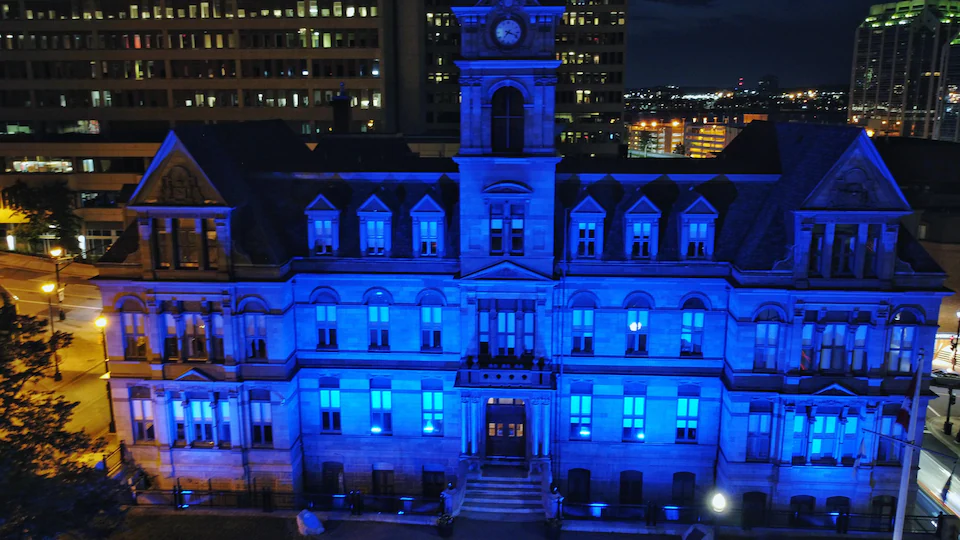 L'hôtel de ville illuminé en bleue et de hauts édifices à l'arrière, de nuit, le tout filmé par un drone.