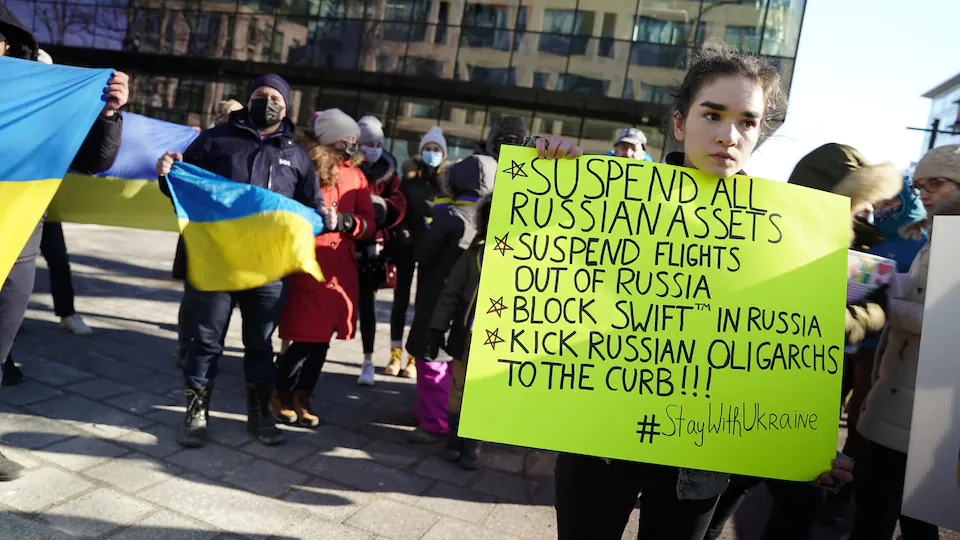 A young woman standing in a public square holds a yellow placard with a list of sanctions she wants imposed on Russia.