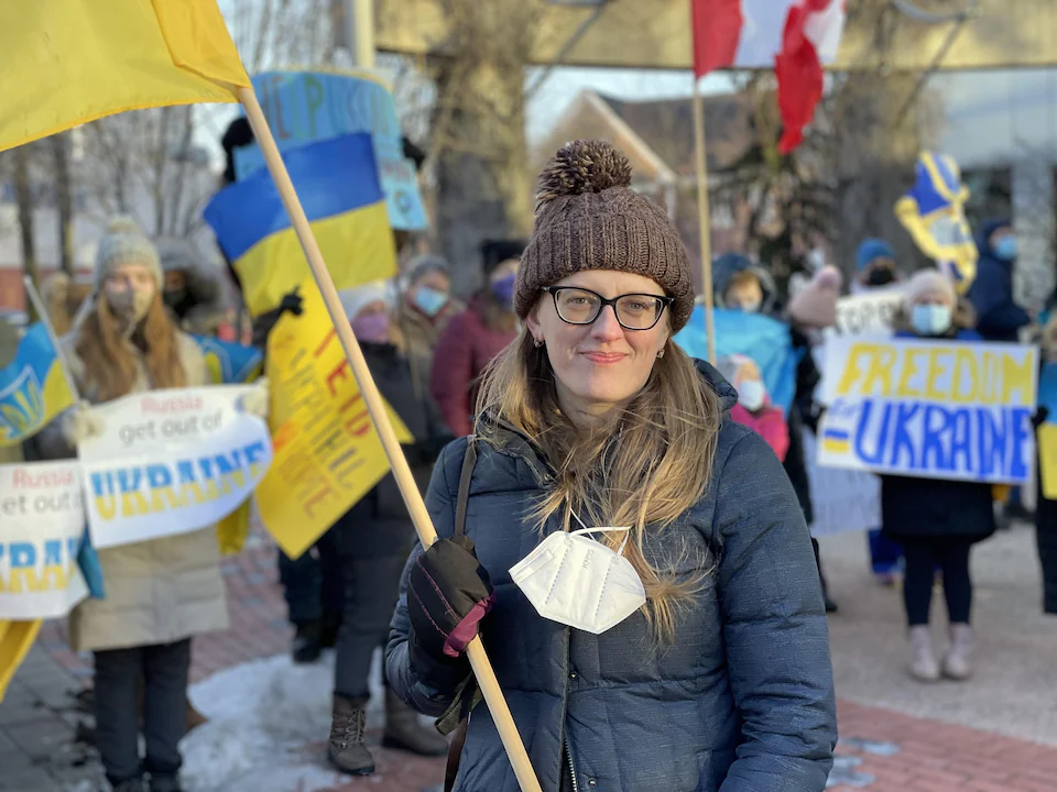 A woman, in the middle of a rally, holds a Ukrainian flag and stares at the camera.