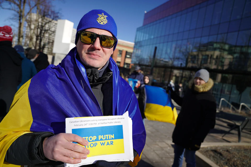 A man outside carries a Ukrainian flag over his shoulders, a blue headdress in his country's colors, and holds a sign that reads. 