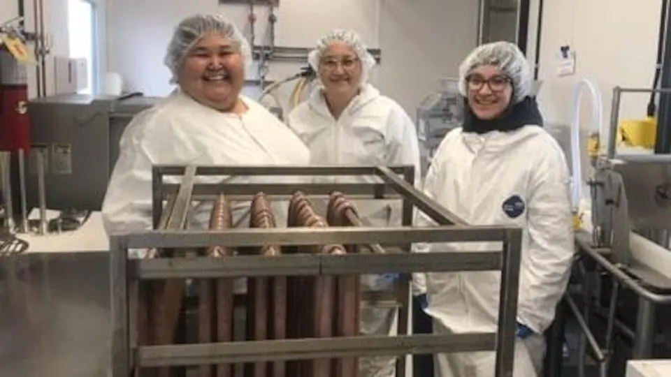 Trois femmes portant des vêtements de protection posent dans une pièce d'usine où sèchent des saucisses.