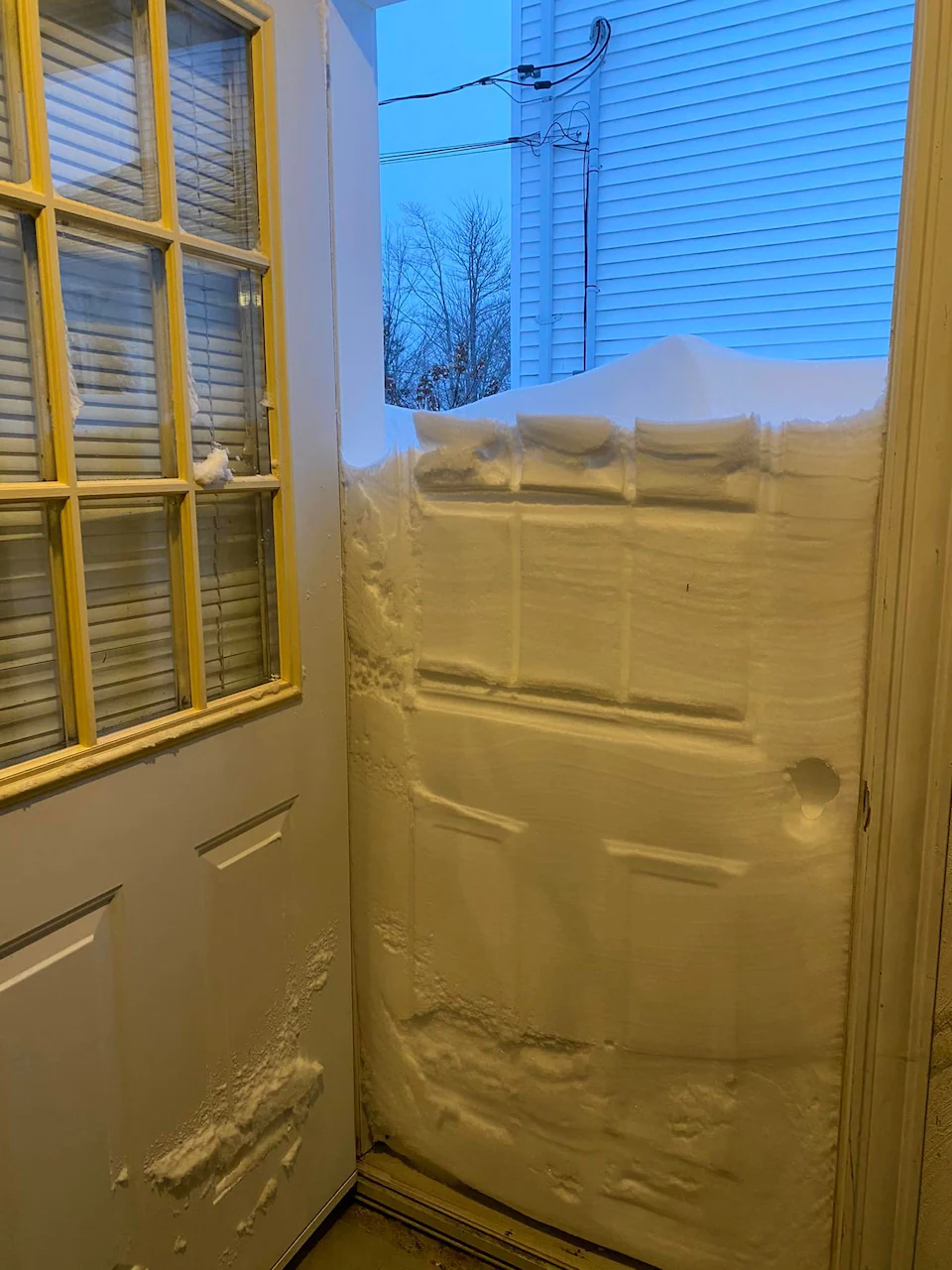 Une porte ouverte laisse voir un demi-mur de neige qui bloque l'accès et empêche de sortir d'une maison.
