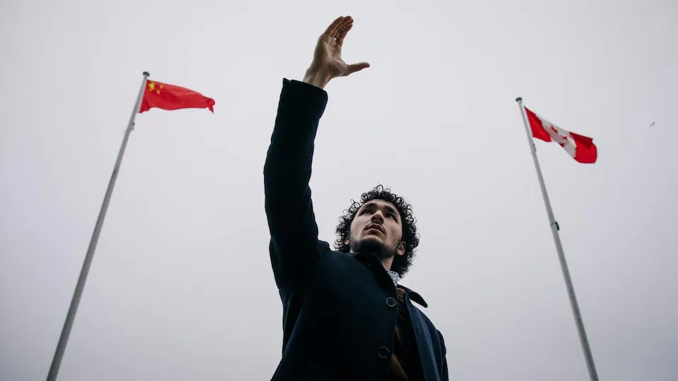 Kabir Qurban lève le bras et fait un signe en forme de croissant avec sa main, devant les drapeaux de la Chine et du Canada.