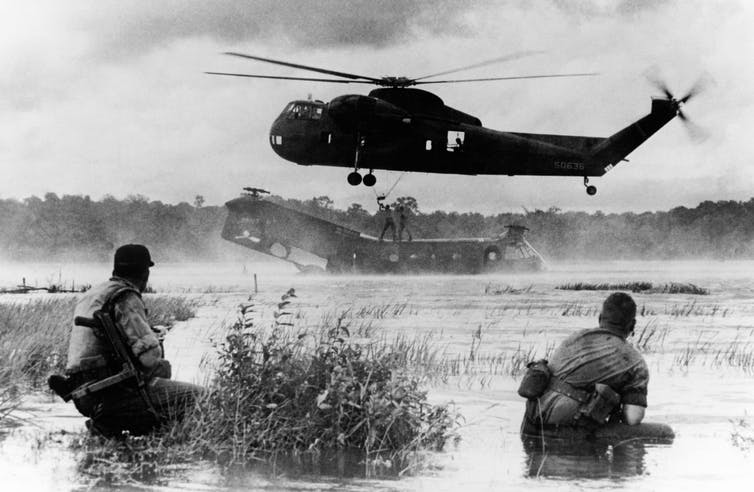 Two soldiers confront a helicopter recovering another helicopter from a body of water in Vietnam in this black and white photo