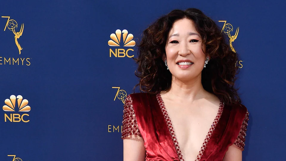 Sandra Oh sourit sur le tapis rouge des prix Emmy devant un mur bleu.