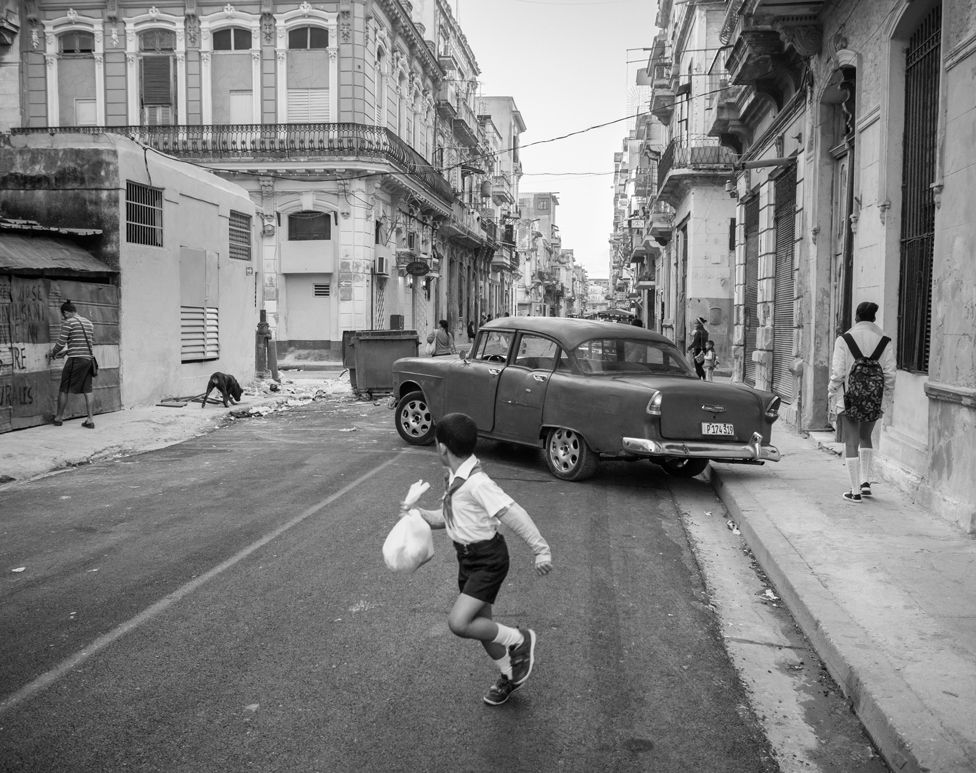 Black and white photo of a child looking behind them at a car as they run in a street