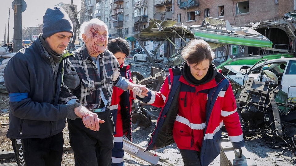 Un homme âgé au visage ensanglanté marche dans des débris, aidé par trois personnes.