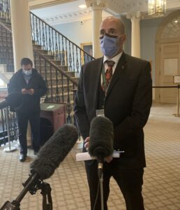 A bald man in a suit and wearing a mask stands at a microphone