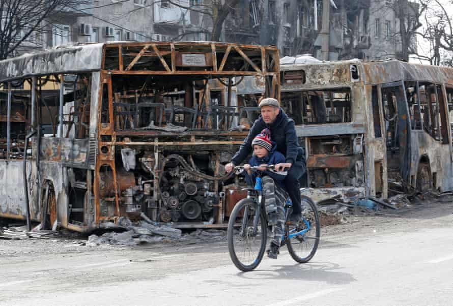 A man and a child ride a bicycle past burnt out buses during Ukraine-Russia conflict in the southern port city of Mariupol, Ukraine.
