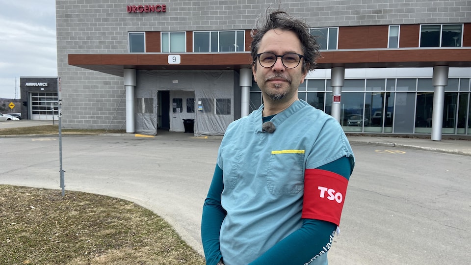 A nurse in a work uniform with a red armband on his left arm stands in front of the emergency room at Montmagny Hospital.