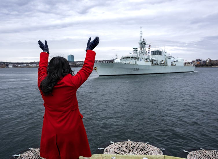 A woman in a red coat with dark hair is seen from behind as she waves at a patrol ship in a harbour.