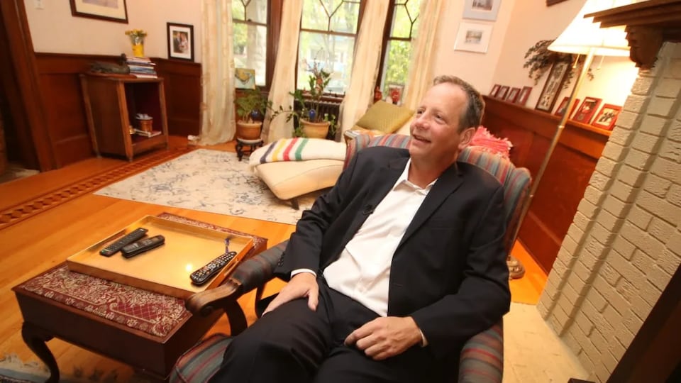 John Orlikow is seated on an armchair in the living room of a house.