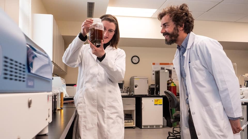 Taylor Belansky and Guillaume Nielsen examining a jar filled with a thick liquid substance in their laboratory.