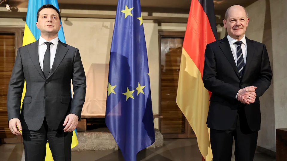 Volodymyr Zelensky et Olaf Scholz debout devant des drapeaux de l'Ukraine, de l'Union européenne et de l'Allemagne.
