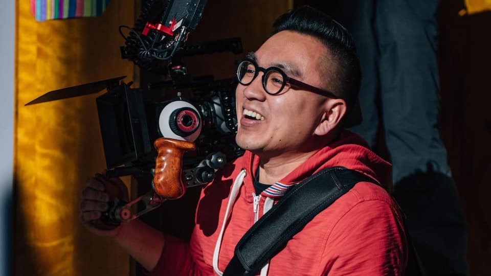 A director with a camera on his shoulder, who smiles.