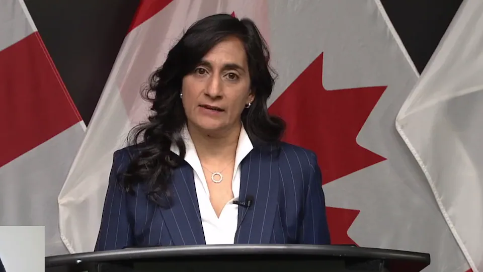 Anita Anand, devant un lutrin, prend la parole avec en arrière-plan plusieurs drapeaux canadiens 