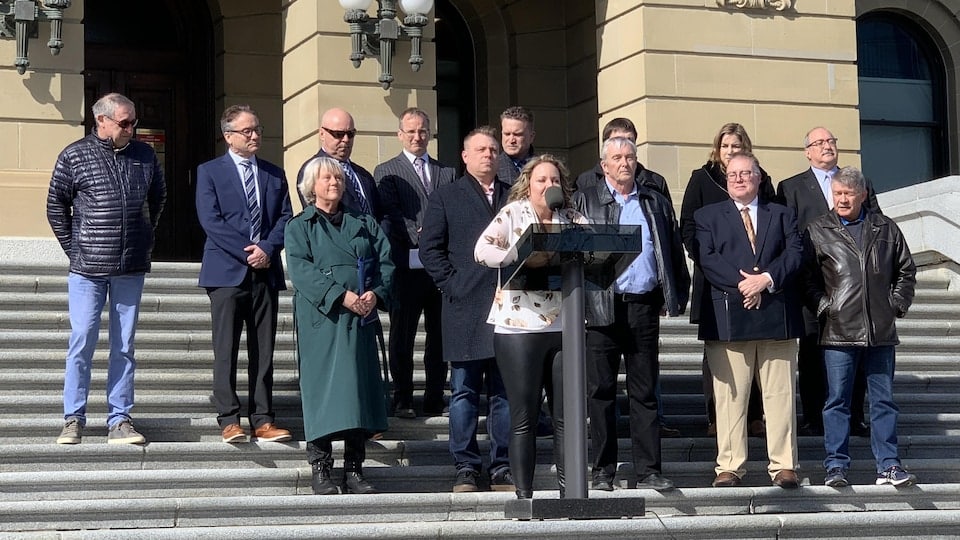 Fourteen people stand on the steps of the Alberta Legislative Assembly, in front of a podium.