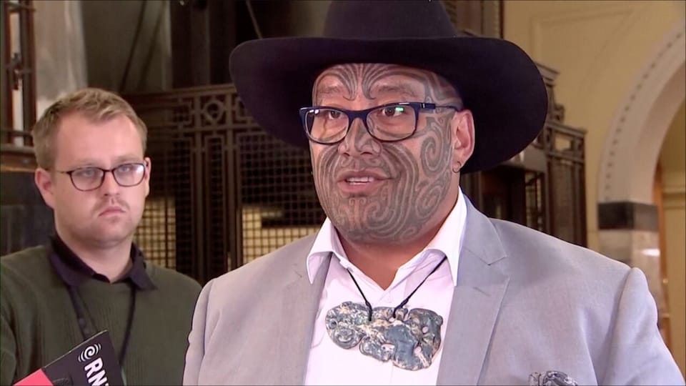 A Maori man has traditional face tattoos and wears glasses and a black hat.