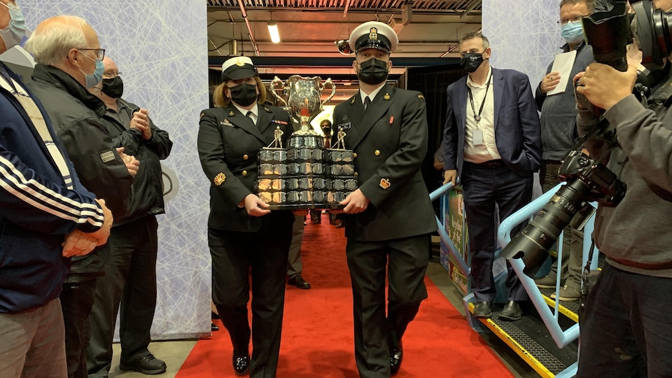 The Memorial Cup is coming to St-Jean.  Police officers carry the Cup, in front of a crowd of people.
