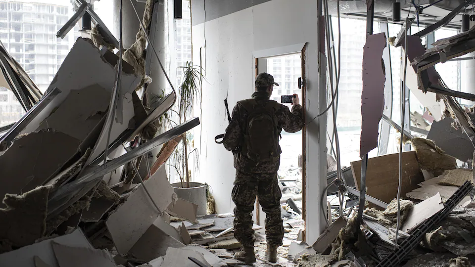 Un homme en habit militaire prend une photo avec son téléphone avec sa main droite. Dans sa main gauche, il tient une arme.