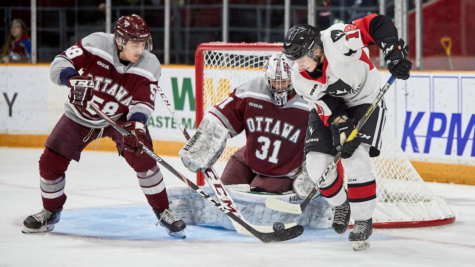 A hockey player in white tries to take a throwback in front of his opponents' net, in gray and garnet, during a college game. 