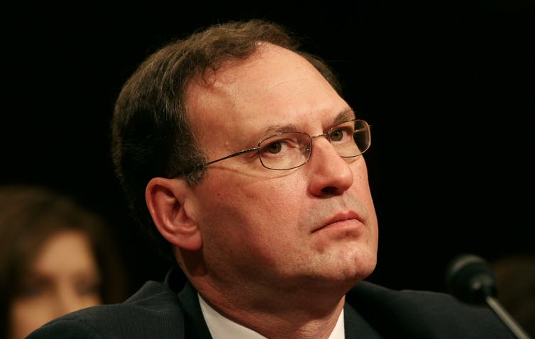 A dark-haired man in a suit, with wire glasses, looking thoughtful.