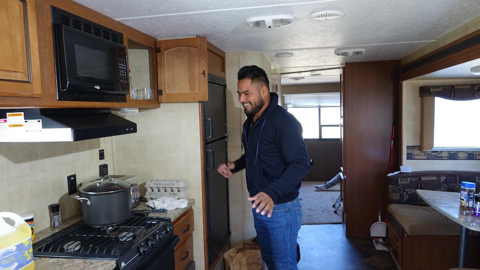 A smiling man stands in the kitchen of the trailer he lives in.