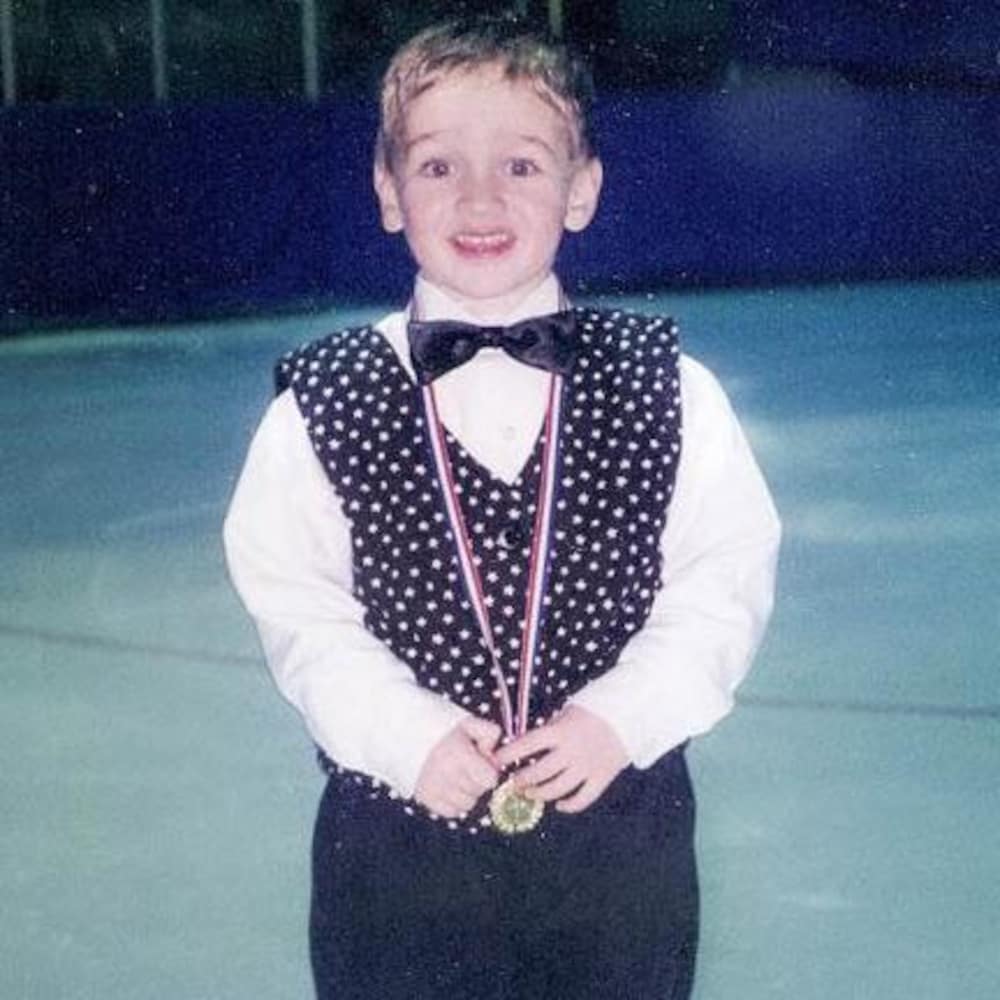 Un jeune garçon se tient debout sur une patinoire et sourit, médaille au cou.