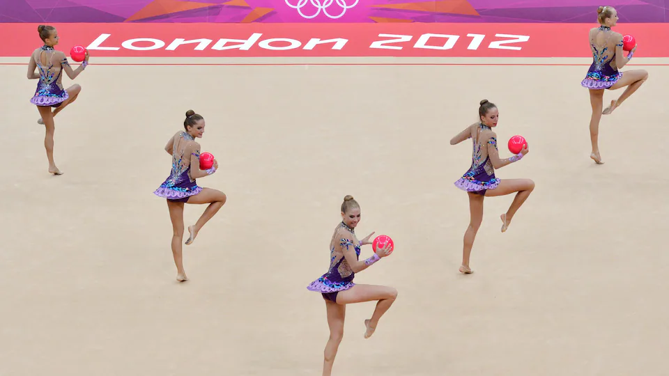 Cinq gymnastes en mauve sur un tapis. Elle tiennent chacune un ballon rose dans leurs mains. 