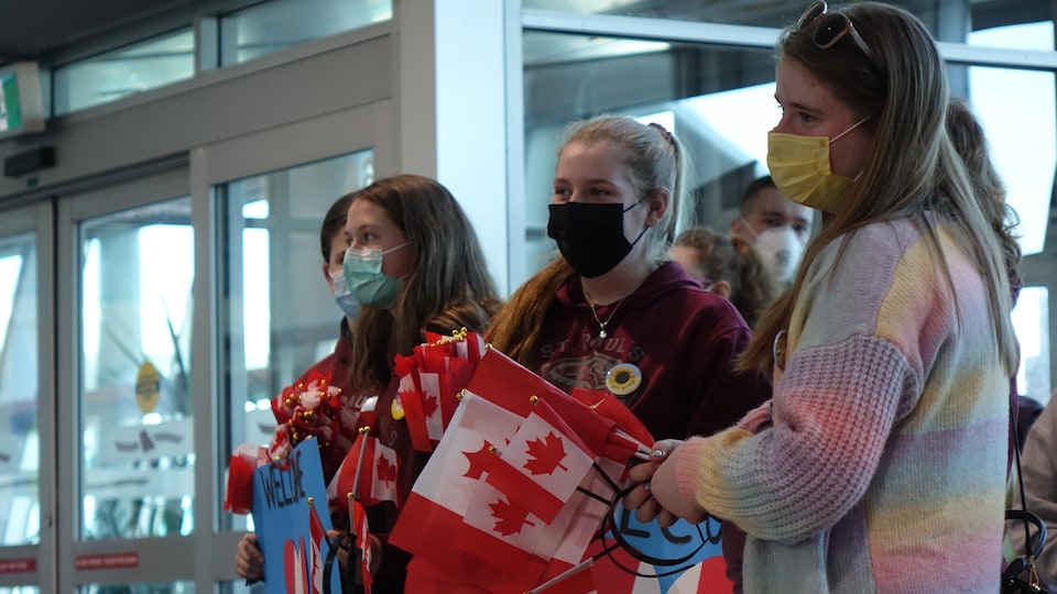 Teenage girls wearing sanitary masks and with Canadian flags in their hands are at an airport. 
