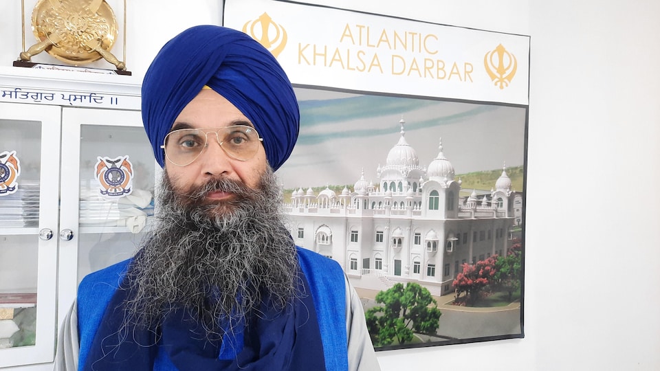 Man with big beard and blue turban in front of photo of Sikh temple.