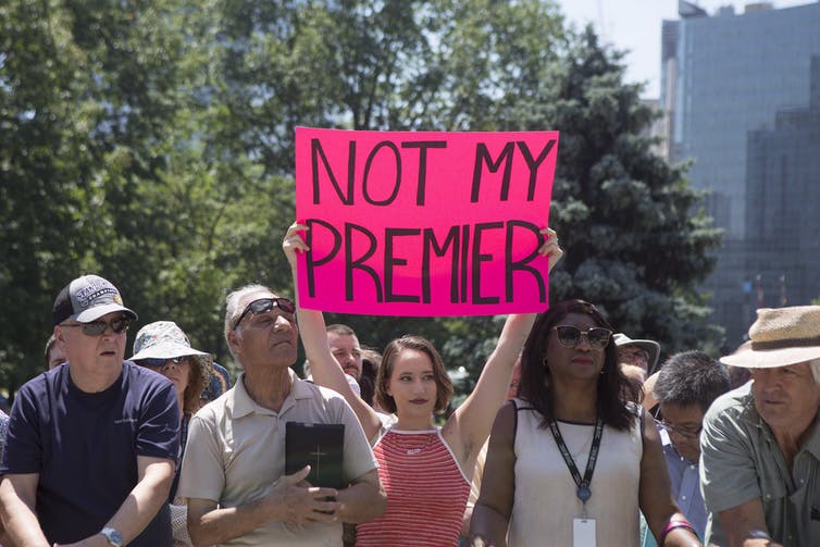 A woman in a sleeveless red-and-white shirt holds up a sign that reads Not My Premier.