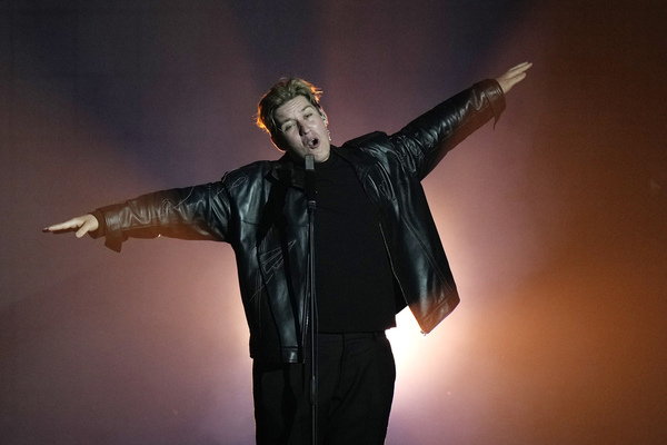A male singer in a black shirt, black pants and a black leather jacket stands with open arms singing on the Eurovision stage.