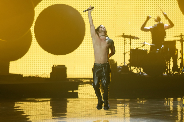 A bare-chested lead singer in black leather pants jumps onto the stage at Eurovision with yellow lights, drums and dark balloons in the background.