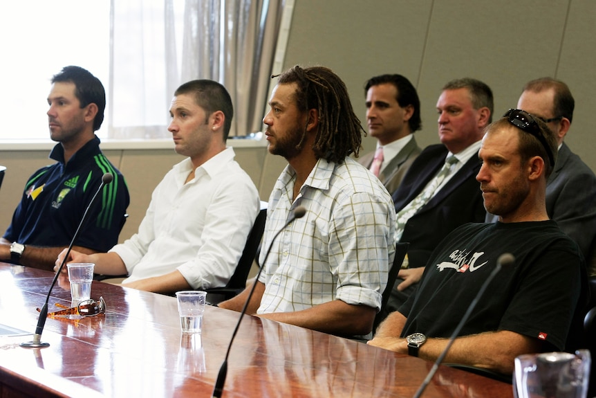 A group of men sit at a long desk with microphones.