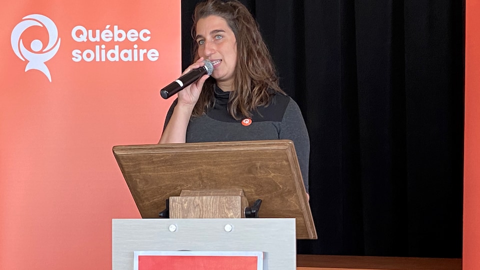 A woman with a microphone in her hand behind a desk on which is written Québec solidaire 