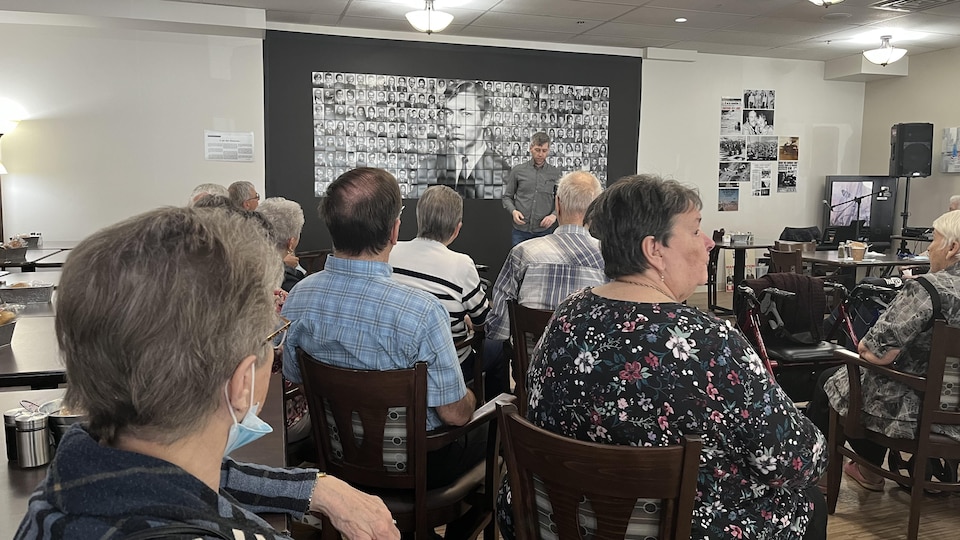 A dozen people seated attend David Simard's speech.  At the back, a banner displays the face of Herman St-Gelais and the 35 other people injured during the attack of May 10, 1972.