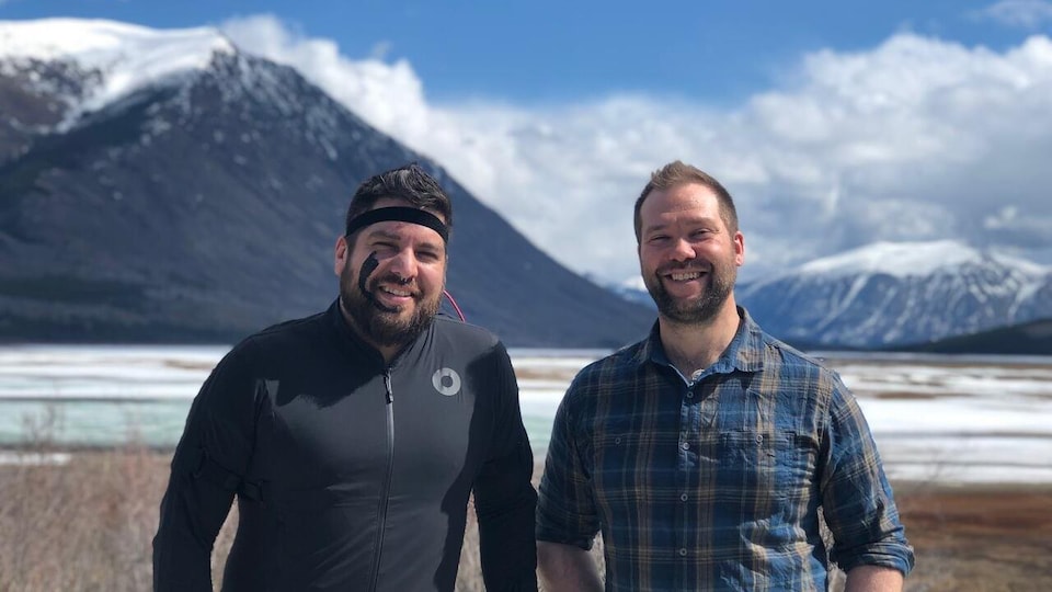 Two men stand in front of a mountain landscape and smile at the camera.  The man on the left is wearing a black motion capture suit.