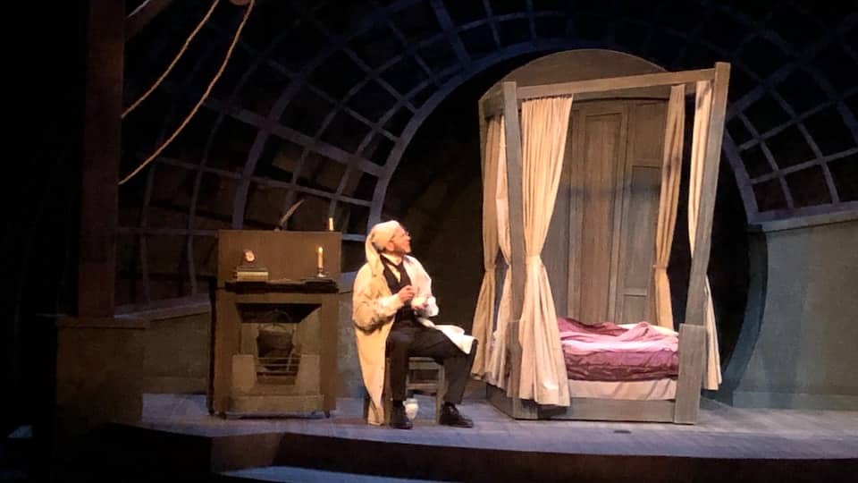 A comedian in the role of Ebenezer Scrooge, seated in a miser's bedroom setting. 