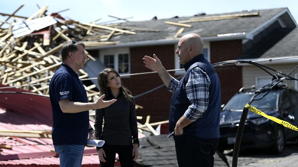 Three politicians in discussion in front of the damage caused by a storm.