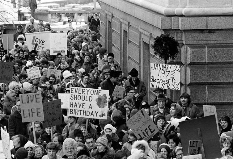 A crowd of men and women protesting near a city building in the winter, holding signs that say 'Love life' and 'Everyone should have a birthday.'