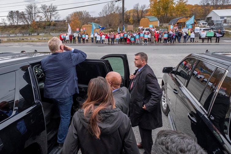 A man gets into a car with as a line of protesters standing in a row across the street holding signs.