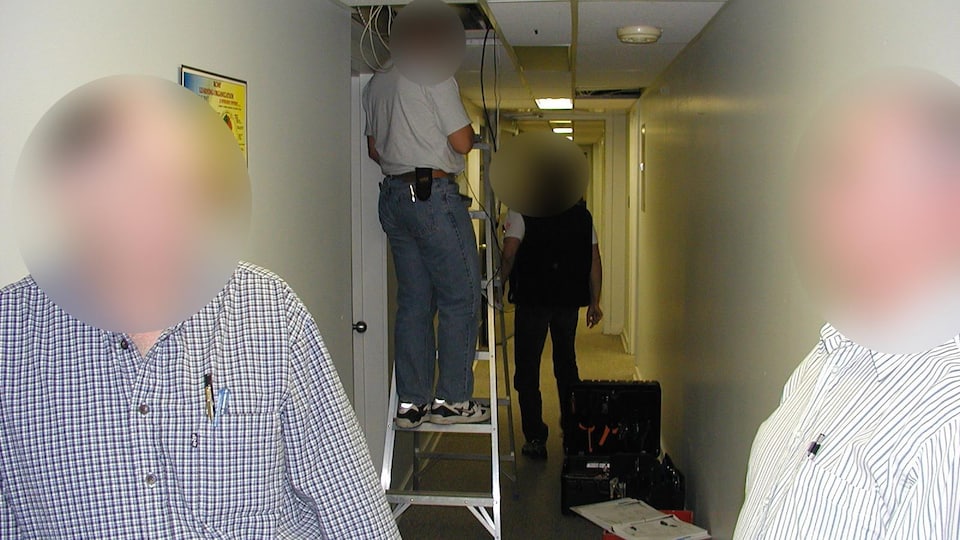 People with blurred faces are in a hallway.  One of them is on a stepladder and is holding cables coming out of the ceiling.
