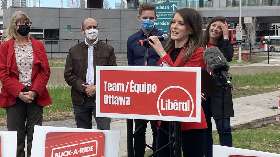 Behind an Ontario Liberal Party sign, Amanda Simard speaks on the microphone with people in the background.