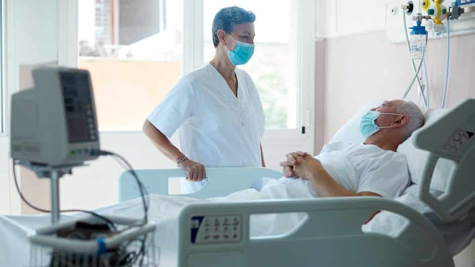A masked nurse talks to a bedridden patient in a hospital room. 